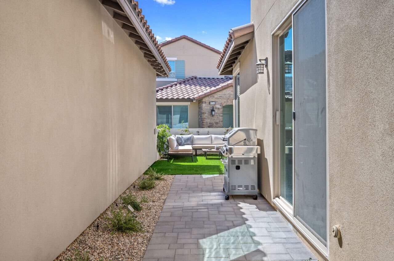 A narrow outdoor patio with a grill, a sofa, and a small lawn area.