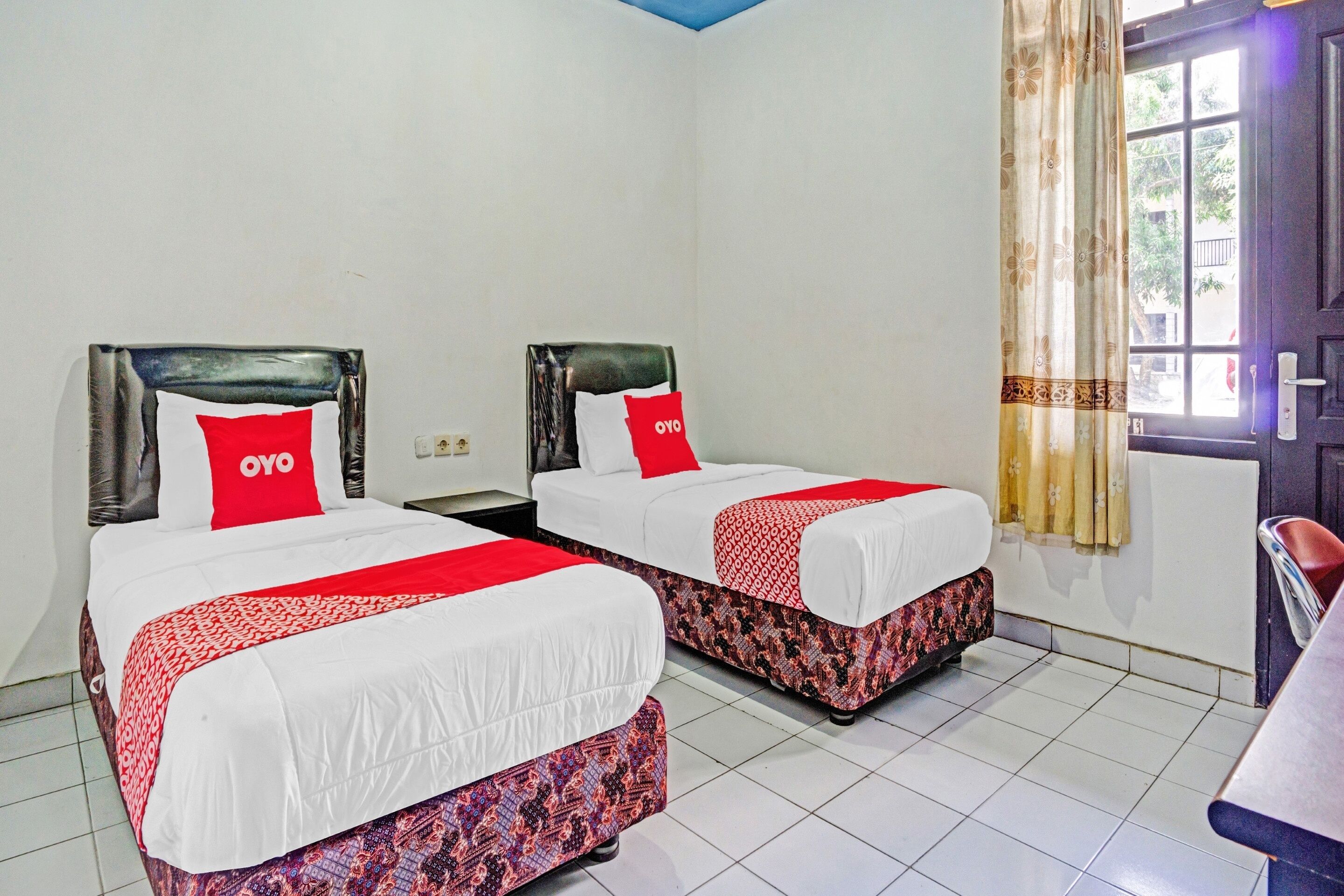 Ein Hotelzimmer mit zwei Betten, jeweils mit einer roten und weißen Tagesdecke und passenden Kissen. Der Raum hat einen Fliesenboden und ein Fenster mit Vorhängen.