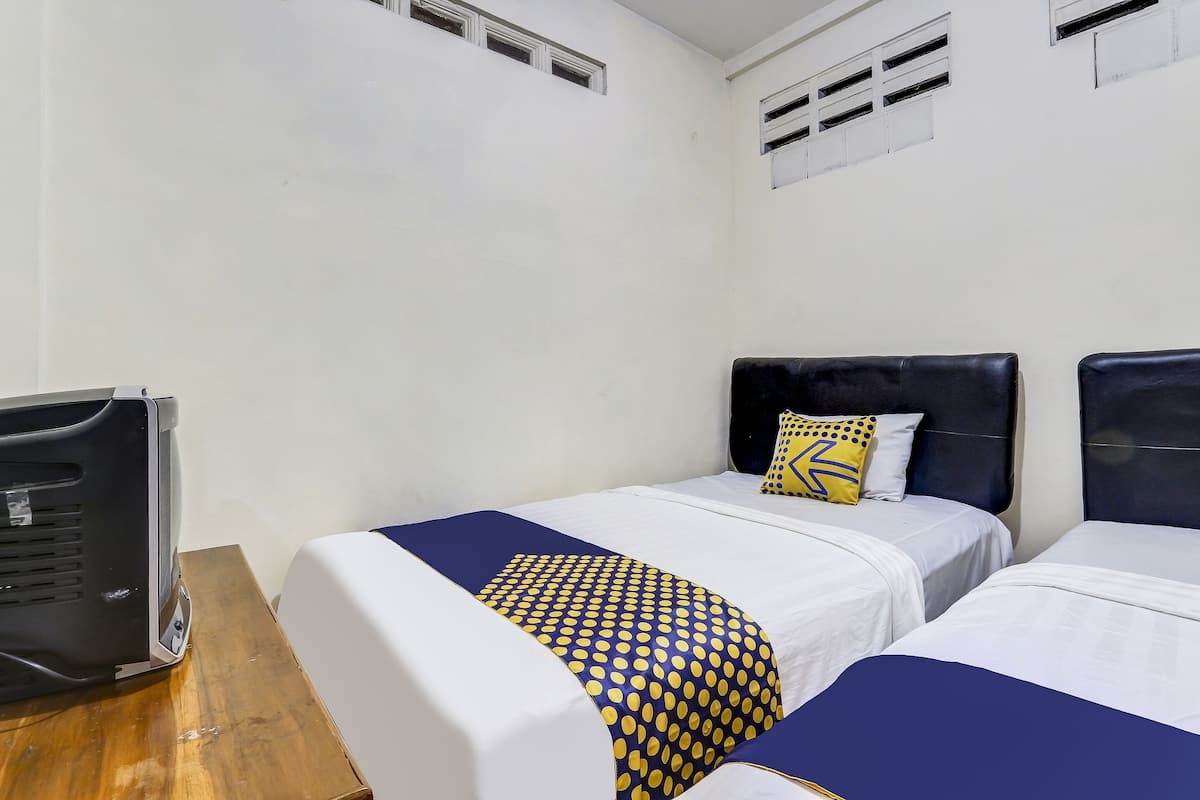 A room with two beds, each with a headboard, white bedding, and a blue and yellow patterned blanket. There is a black speaker on a wooden table.