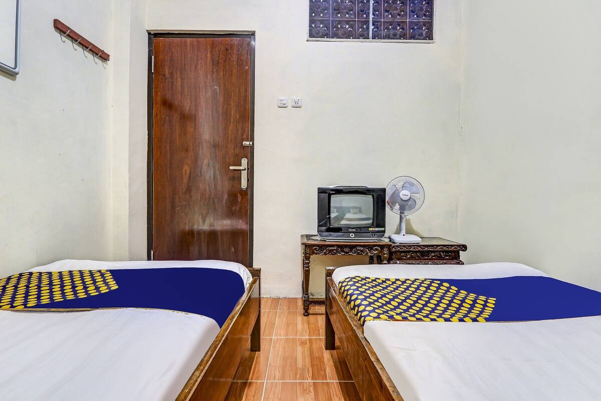 A room with two beds, a wooden door, a small table with a television and fan, and a window with a grid pattern.