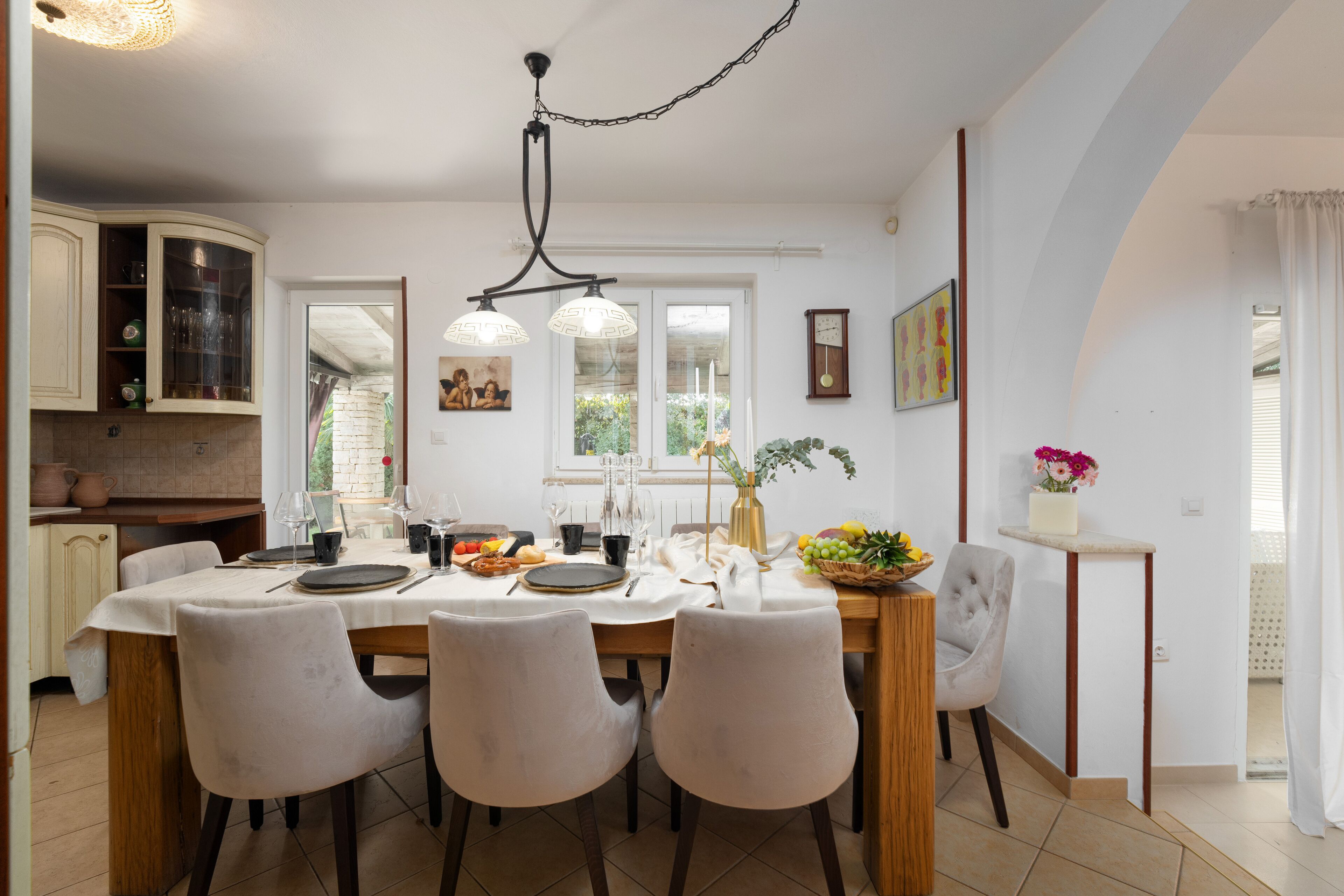A dining area with a wooden table, four chairs, and a chandelier. There is a kitchen with cabinets and a window in the background.