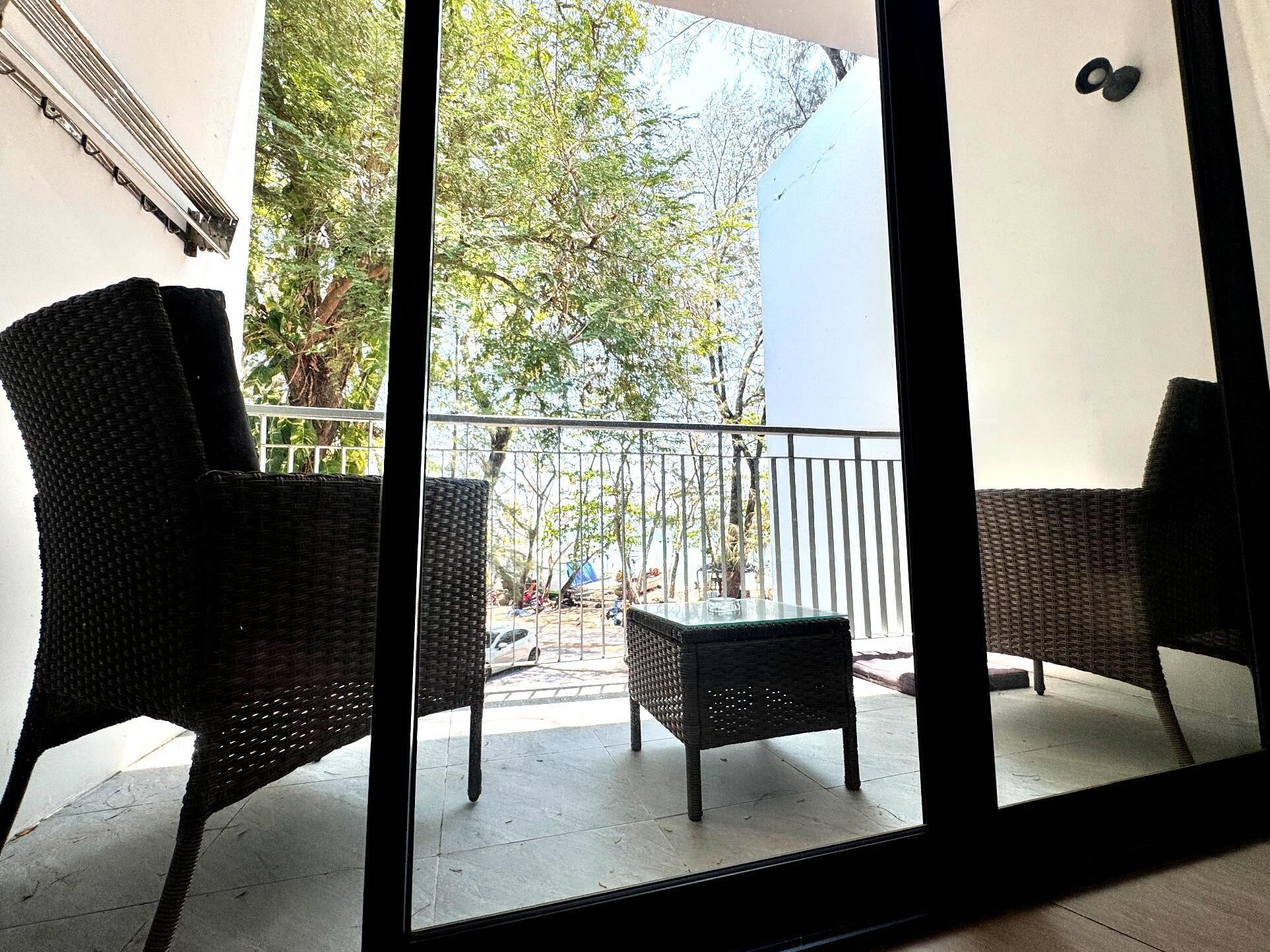 Un balcón con mobiliario de mimbre, una mesa de vidrio y vistas a árboles y una calle.