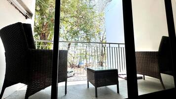 Un balcón con mobiliario de mimbre, una mesa de vidrio y vistas a árboles y una calle.