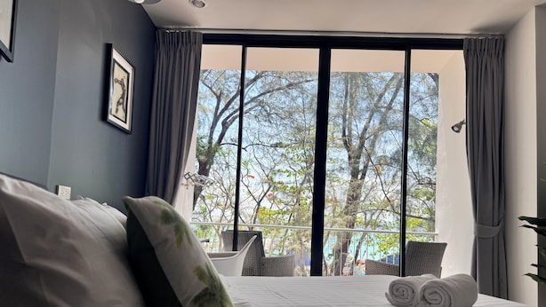 A hotel room with a bed, pillows, a view of trees through a large window, and a balcony with chairs. - Phuket Muay Thai Hotel Rawai Beach (Rawai)