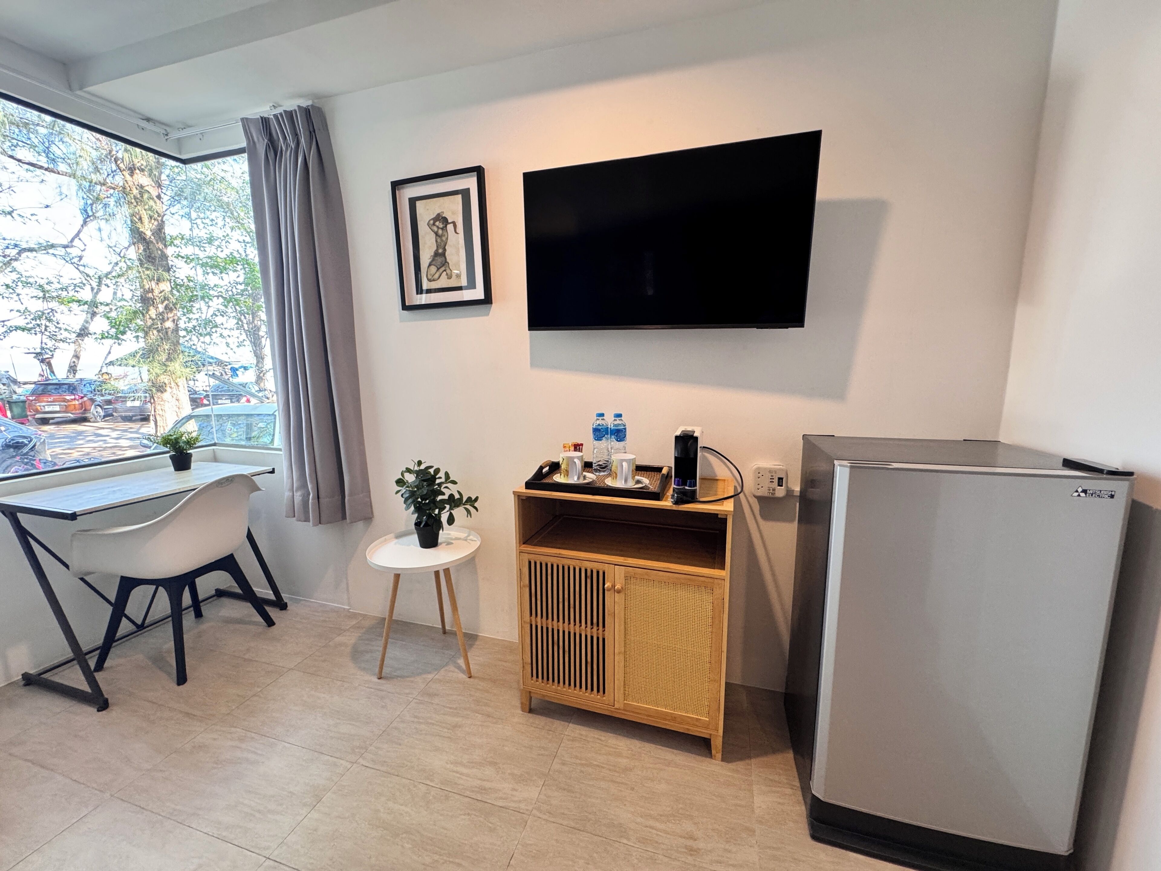 A compact room with a desk, chair, potted plant, small side table, mounted TV, and a refrigerator.