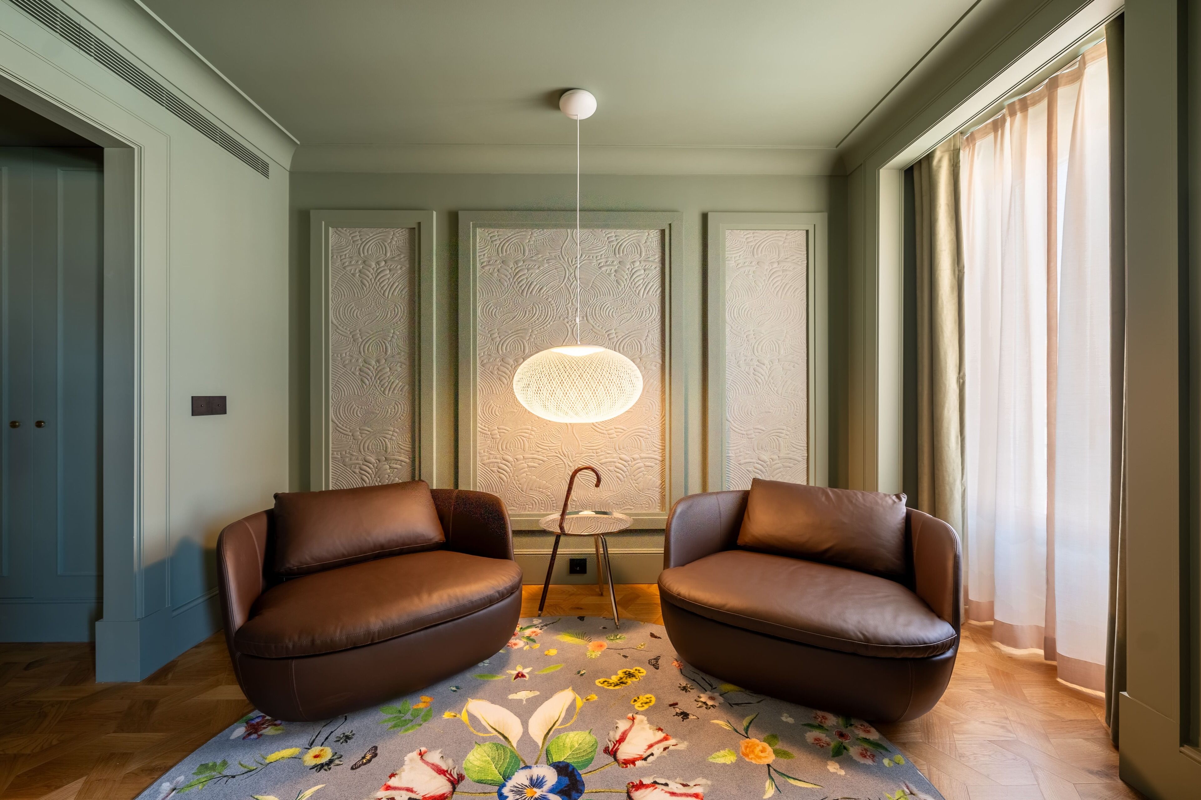 A room with two brown leather armchairs, a patterned rug, a hanging light fixture, and a window with sheer curtains.