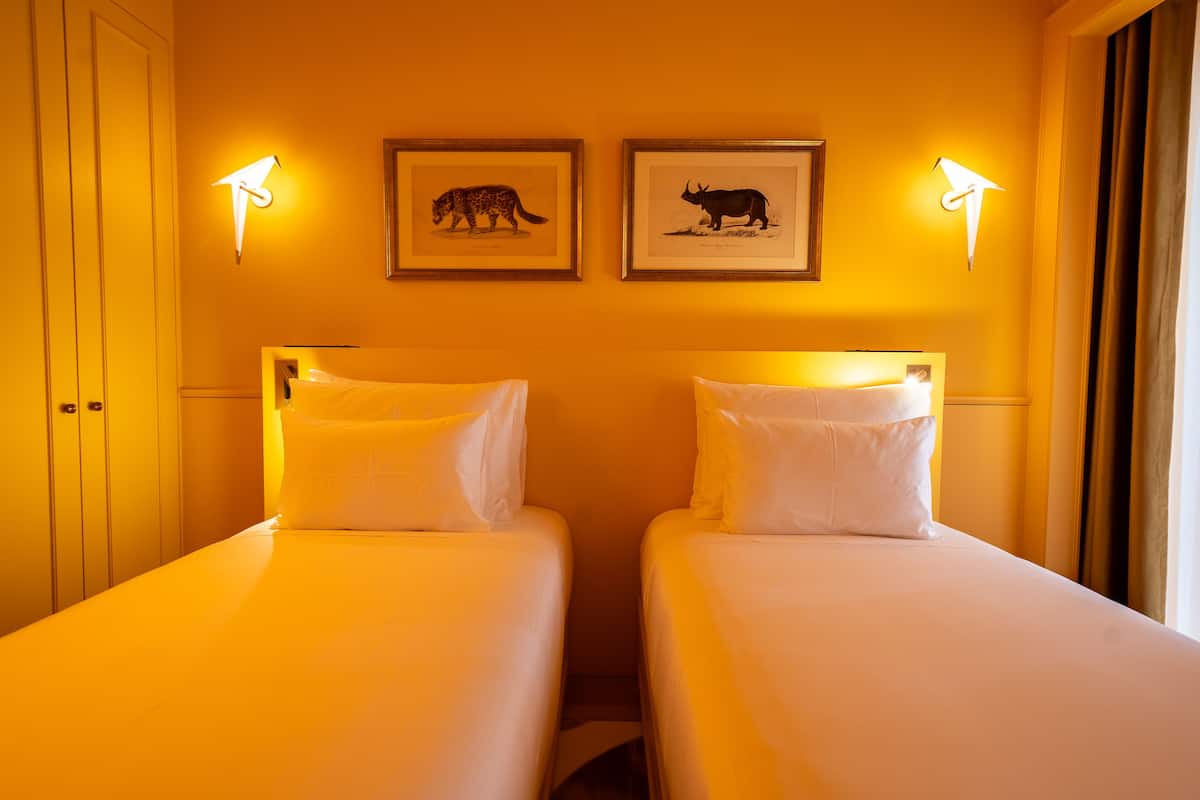Une chambre d’hôtel avec deux lits simples, chacun agrémenté de draps et d’oreillers blancs. Au-dessus des lits, le mur est décoré d’illustrations d’animaux encadrées.