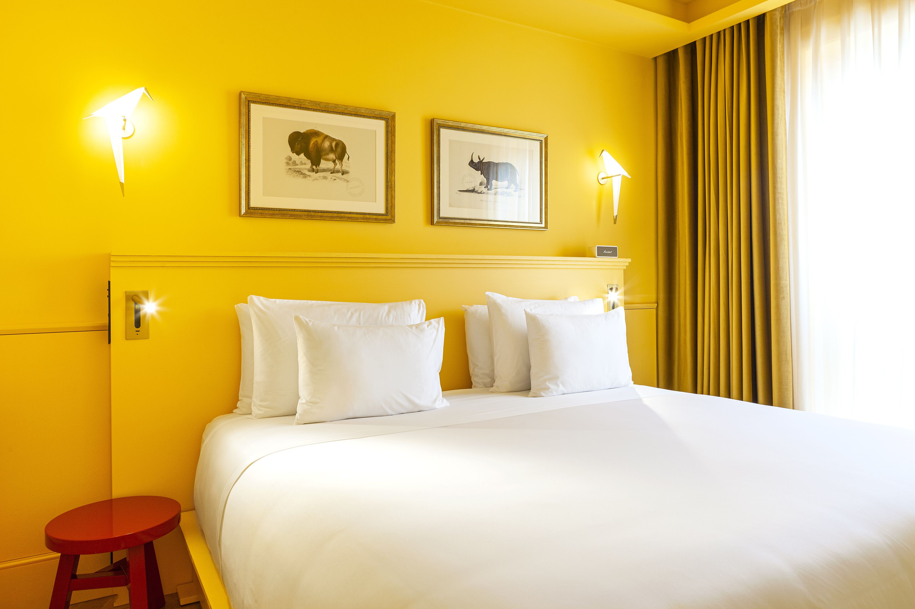 Una habitación de hotel con una cama grande, dos cuadros en la pared y un taburete rojo.