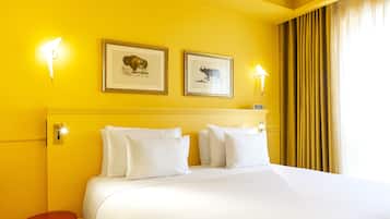 Una habitación de hotel con una cama grande, dos cuadros en la pared y un taburete rojo.