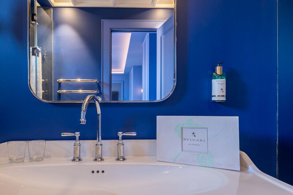 A bathroom with a white sink, two faucets, and a mirror. There is a bottle of BVLGAE and a box of BVLGAE products on the wall.