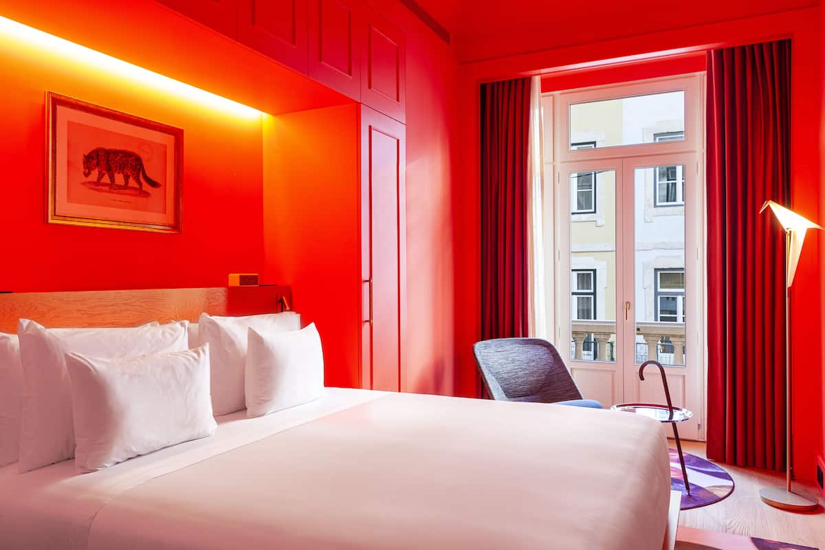 Une chambre d’hôtel avec un grand lit, un tableau représentant un animal, une chaise et une fenêtre avec des rideaux.