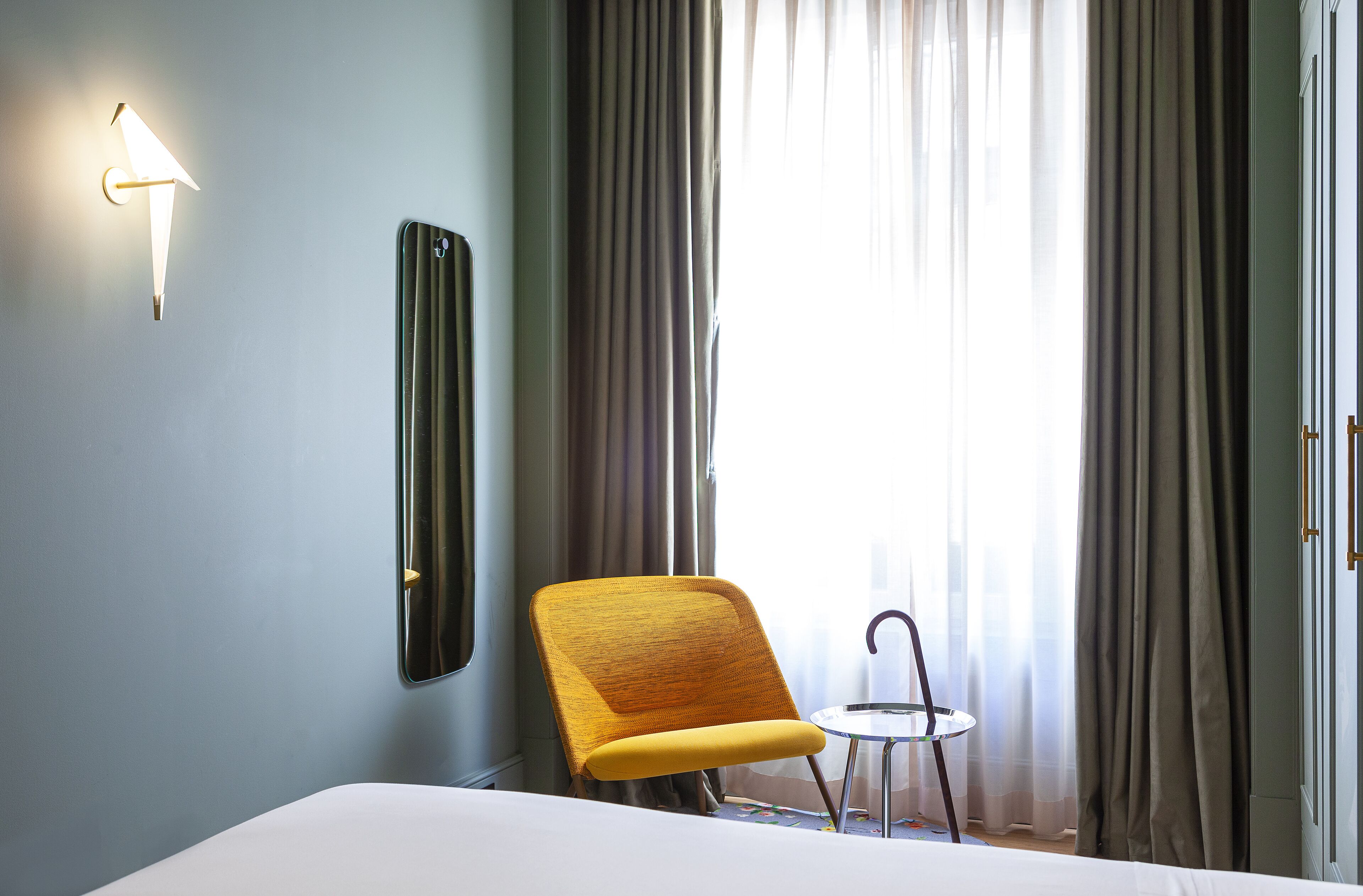 A hotel room with a bed, a chair, a small table, a wall-mounted light, and a window with curtains.