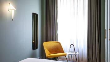 A hotel room with a bed, a chair, a small table, a wall-mounted light, and a window with curtains.