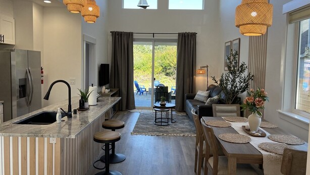 A modern kitchen with a central island, bar stools, and a dining area with a wooden table and chairs. There are two windows with curtains, a sofa, and a plant on the right side. - Drift Inn Duplex B - with hot tub (Ocean Shores)