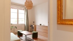 Een moderne kamer met een houten bureau, twee groene poefs, een hangende geweven lamp en een groot raam met gordijnen.