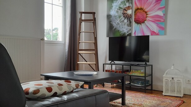 Ein Wohnzimmer mit einem schwarzen Ledersofa, einer Holzlader, einem Flachbildfernseher auf einem schwarzen Standfuß und einem großen floralen Gemälde an der Wand.