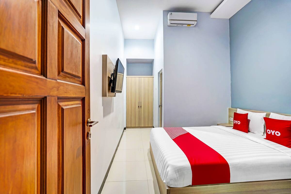 A hotel room with a wooden wardrobe, a bed with red and white bedding, a TV mounted on the wall, and an air conditioning unit.