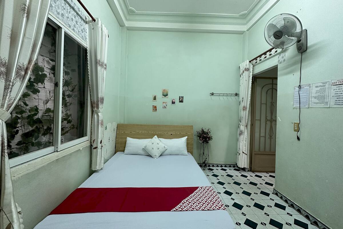 Une chambre à coucher avec un lit, une fenêtre avec des rideaux, un ventilateur fixé au mur et un sol carrelé.