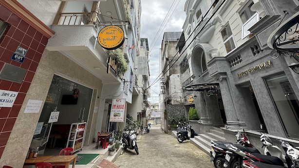 A narrow street with buildings on either side, a sign for a bank, and several parked motorcycles. - OYO 1206 Toan Thang Homestay (Xuan Huong - Da Lat)