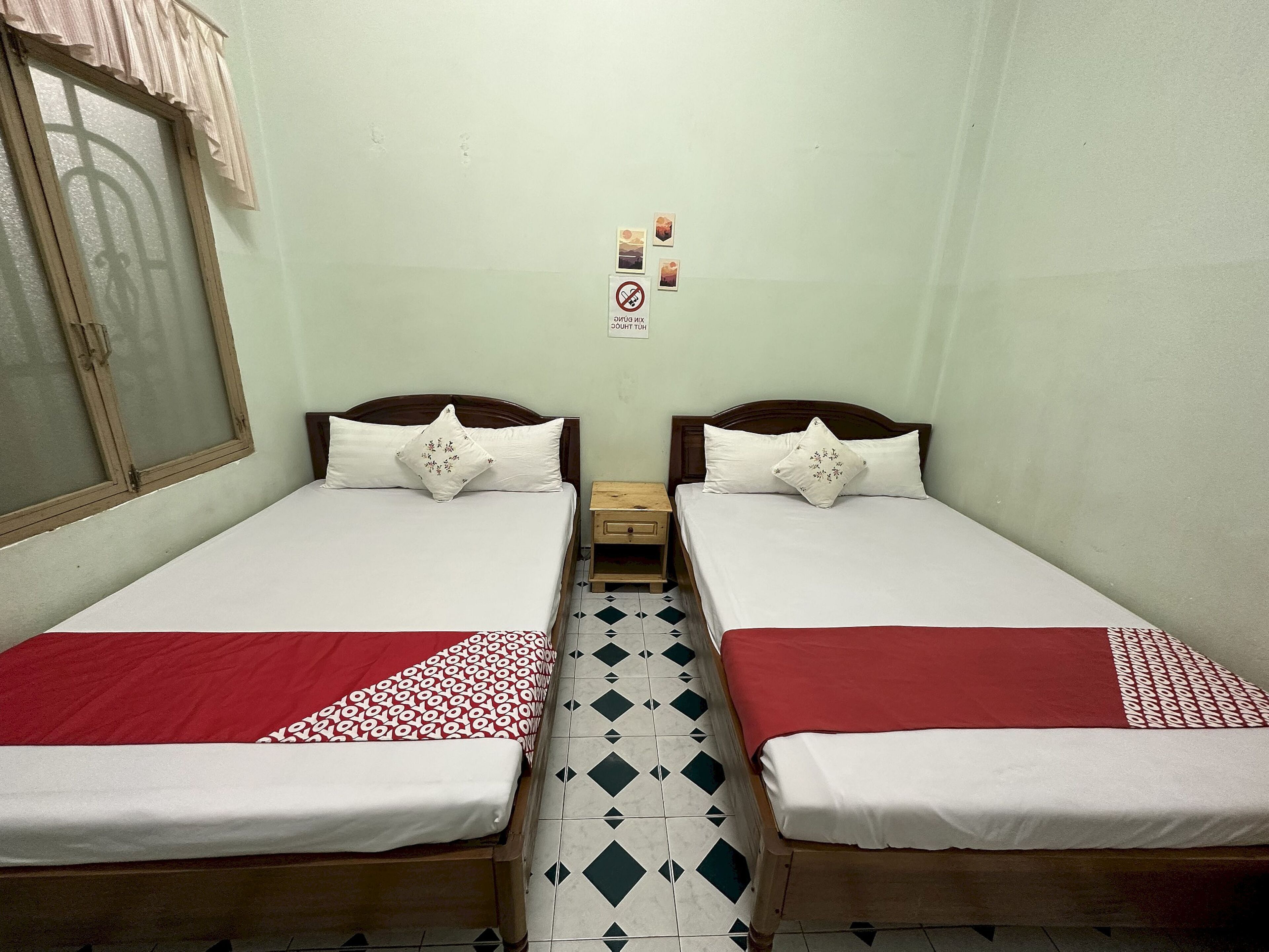 A room with two single beds, each with a red and white bedspread, wooden headboards, and white pillows. There is a small wooden nightstand between the beds, and a window with a wooden frame and curtains. The walls are painted a light color, and there is a small sign with a symbol on the wall.