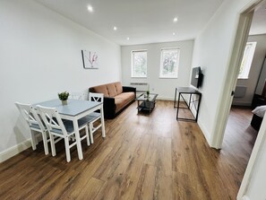 A modern living room with a dining table, white chairs, a brown sofa, and a coffee table. - 1-bed Apartment in Ealing (London)