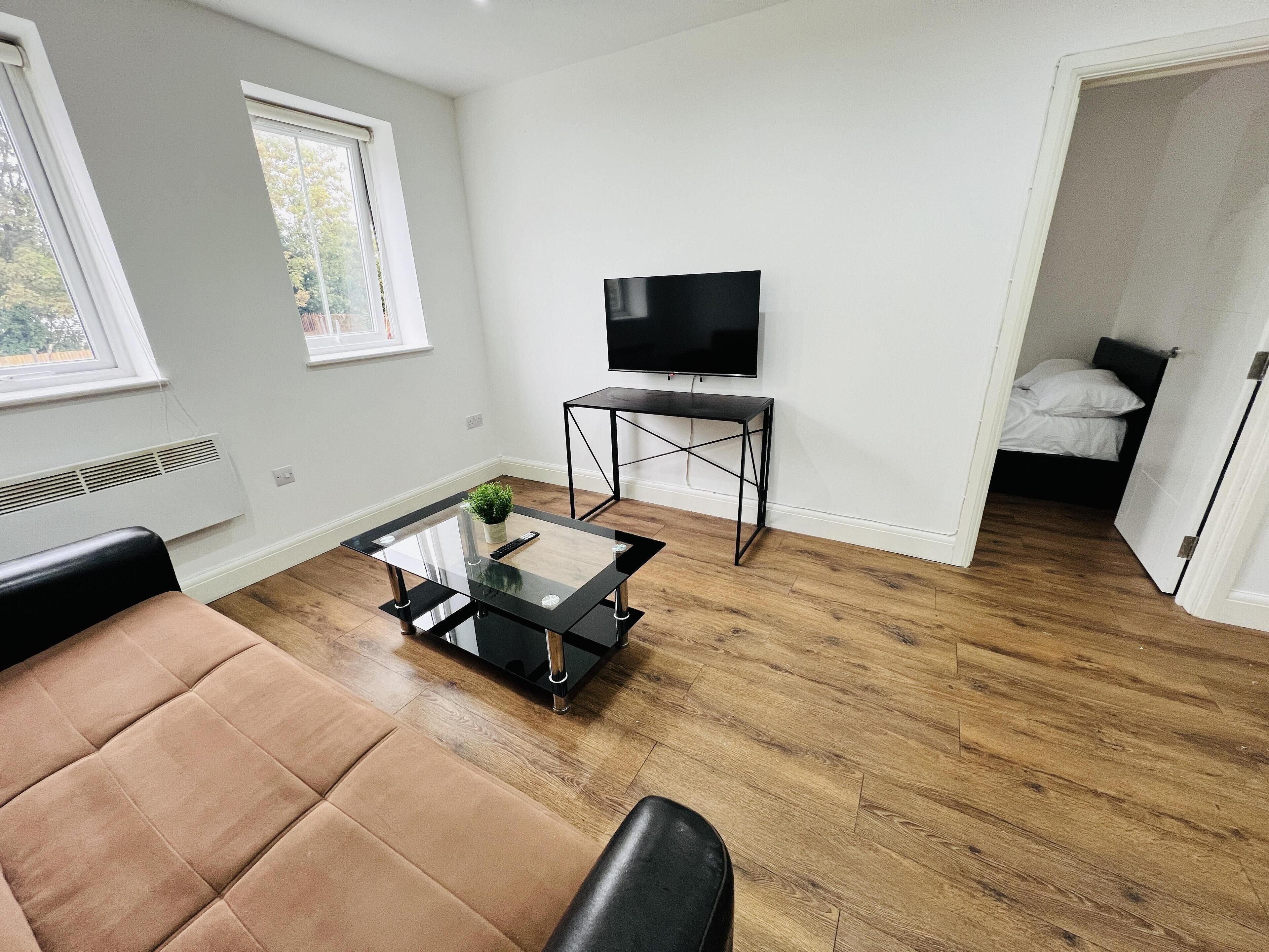 Ett modernt vardagsrum med en lädersoffa, ett soffbord i glas, ett svart TV-bord och en säng i bakgrunden.