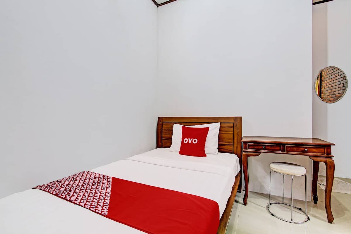 A neatly made bed with a red and white bedspread, a wooden nightstand with a stool, and a wall-mounted round mirror.