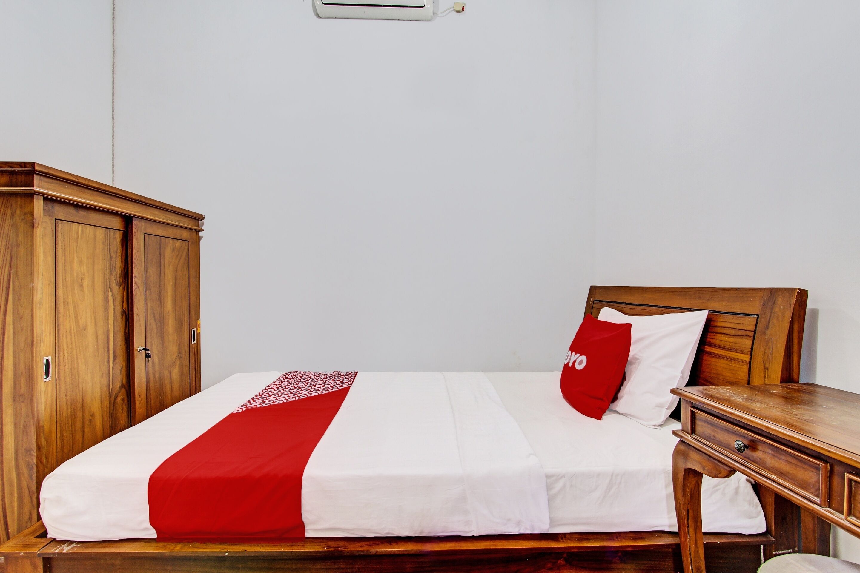 Uma cama bem arrumada com colcha vermelha e branca, uma mesinha de cabeceira de madeira e um guarda-roupa.