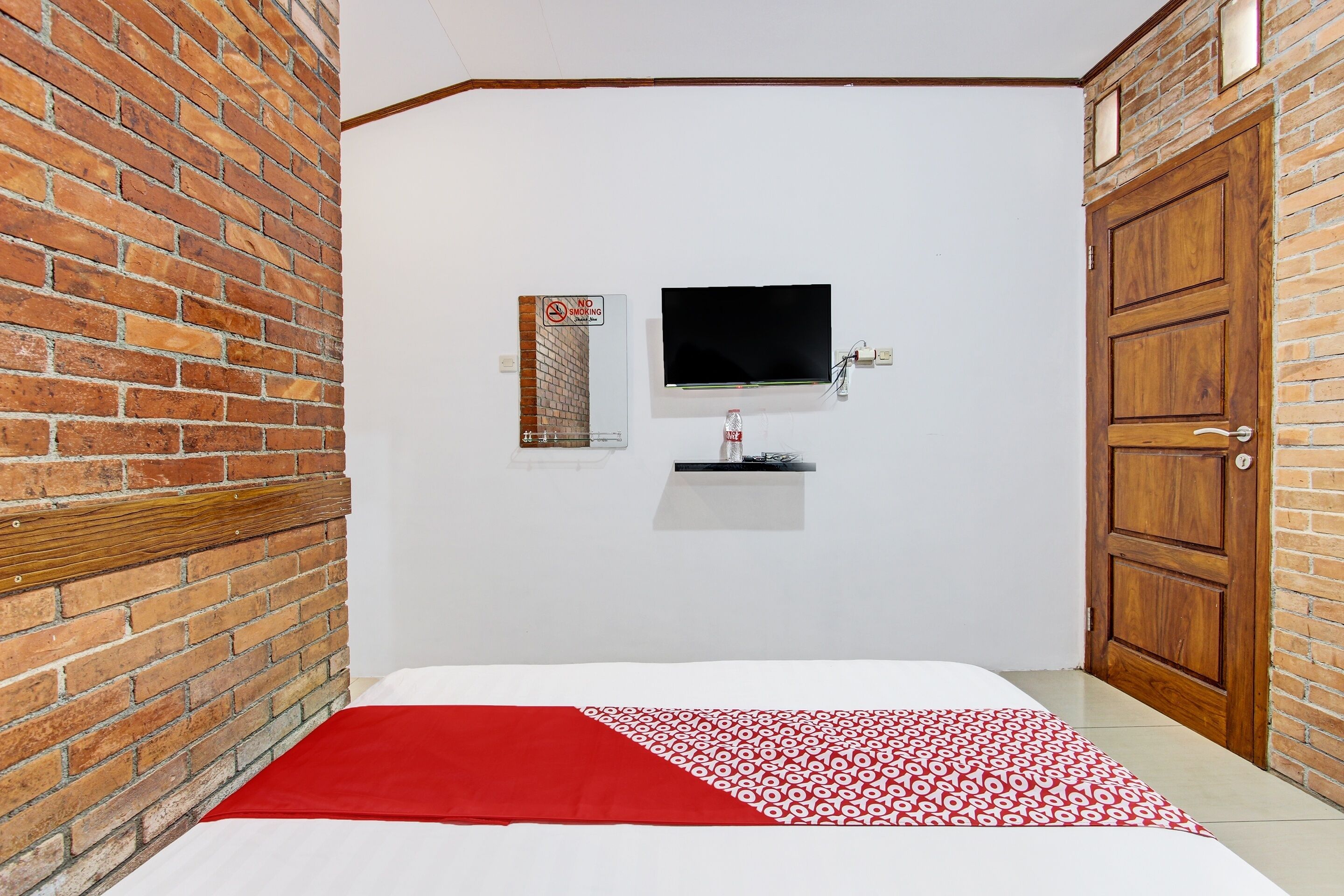 A room with a brick wall, a television mounted on the wall, a wooden door, and a bed with a red and white patterned bedspread.