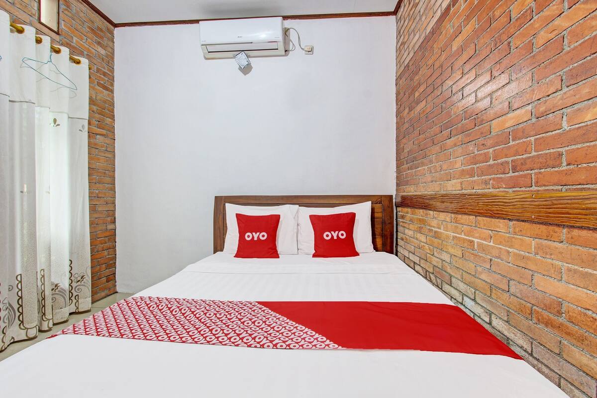 A hotel room with a brick wall, a bed with red and white bedding, and a white air conditioning unit on the wall.