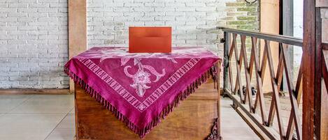 A wooden table with a pink patterned tablecloth and a red object on top, set against a white brick wall with a metal railing in the background.