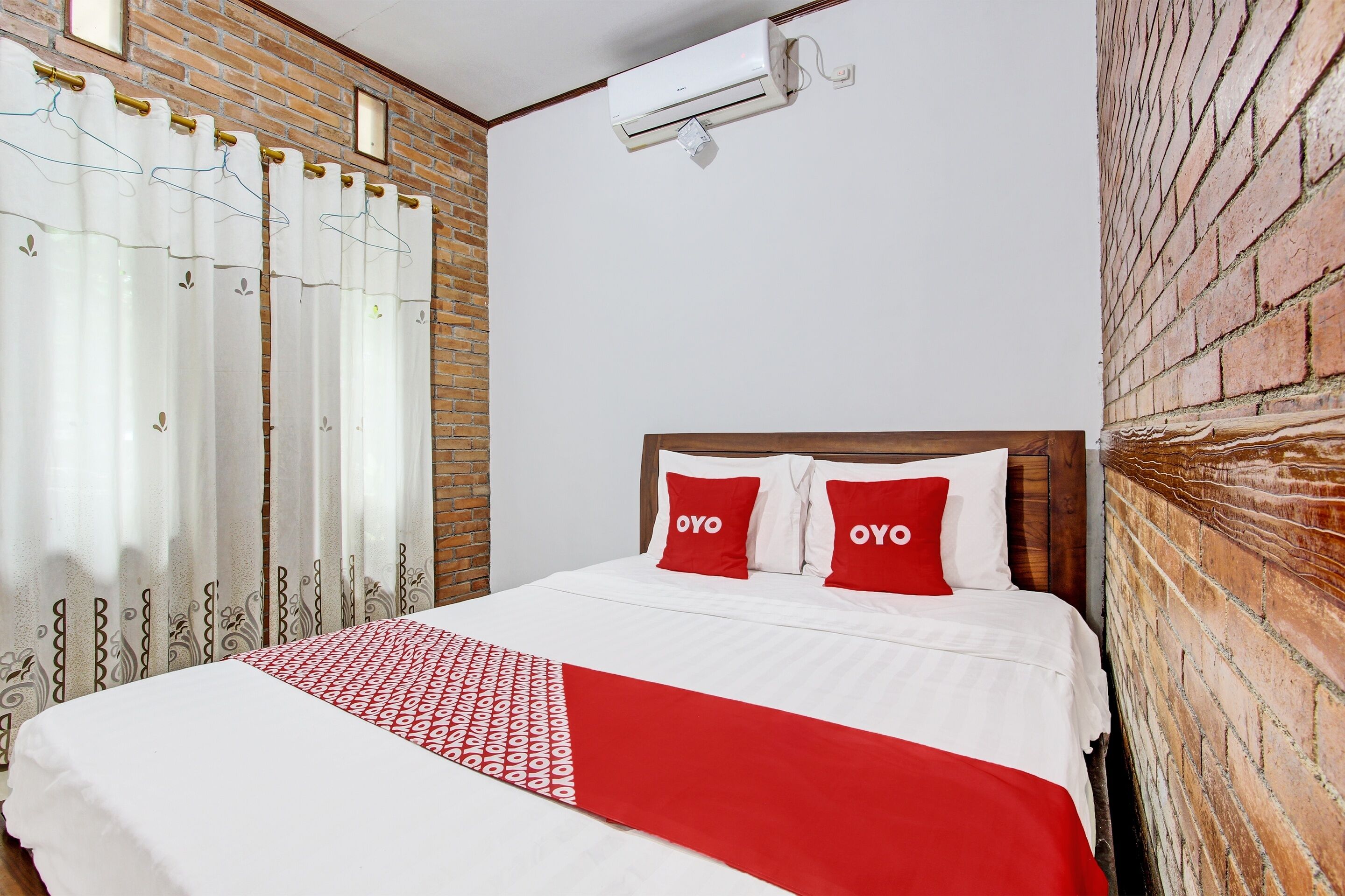 A hotel room with a brick wall, a bed with white bedding and red accents, and a window with sheer curtains.