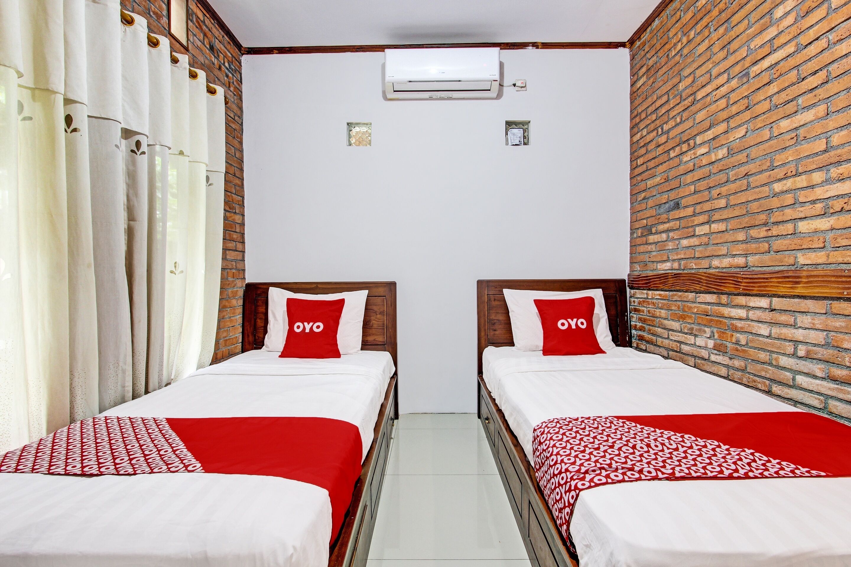Habitación de hotel con dos camas, cada una con cubrecama rojo y blanco y un cojín con la palabra "OYO". Se aprecia una pared de ladrillo en un lado y una pared blanca en el otro.