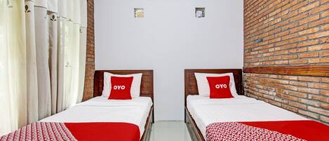 A hotel room with two beds, each with a red and white bedspread and a pillow with the word "OYO". There is a brick wall on one side and a white wall on the other.