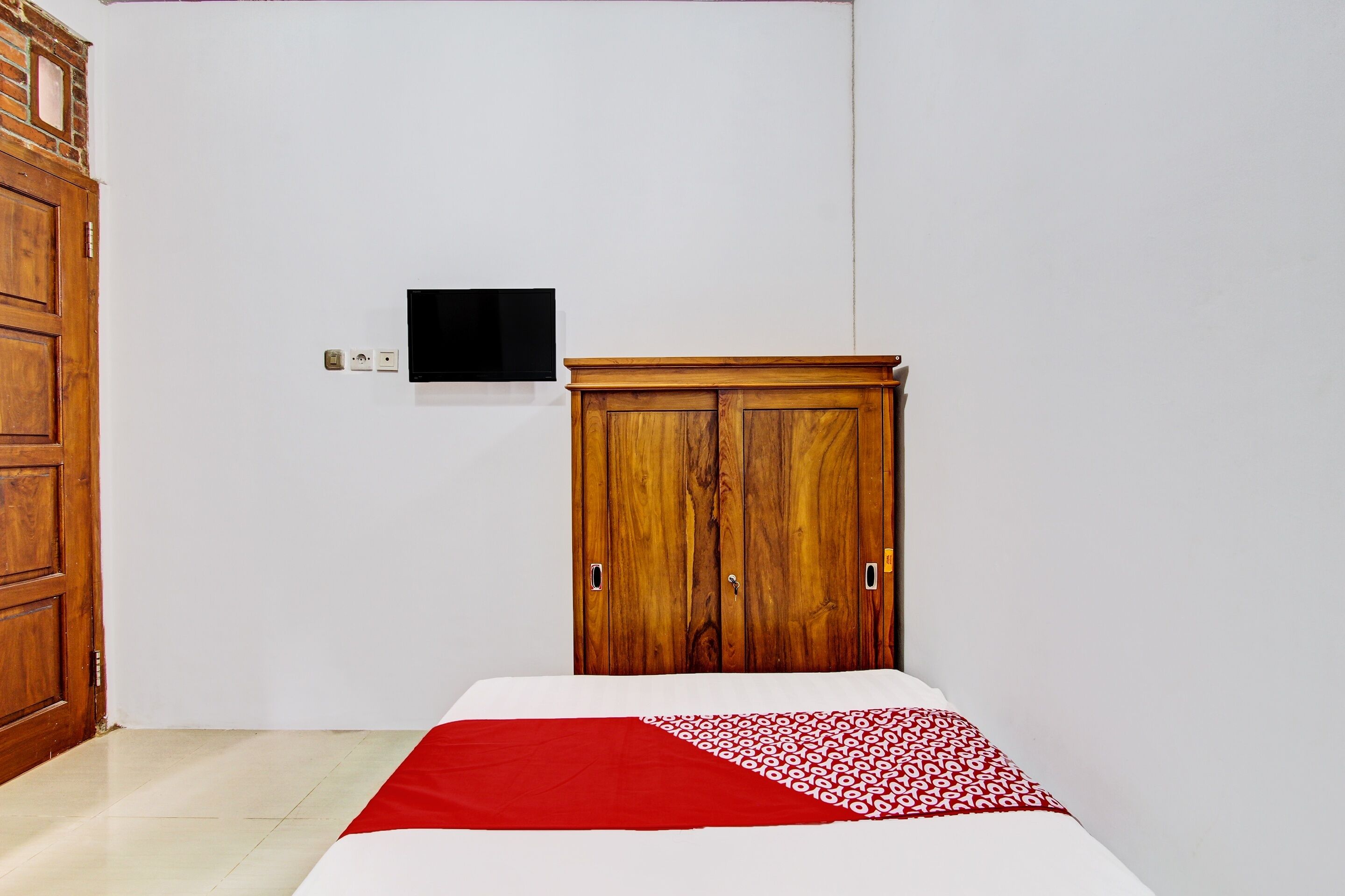 A bedroom with a wooden wardrobe, a bed with a red and white patterned bedspread, and a flat-screen TV mounted on the wall.