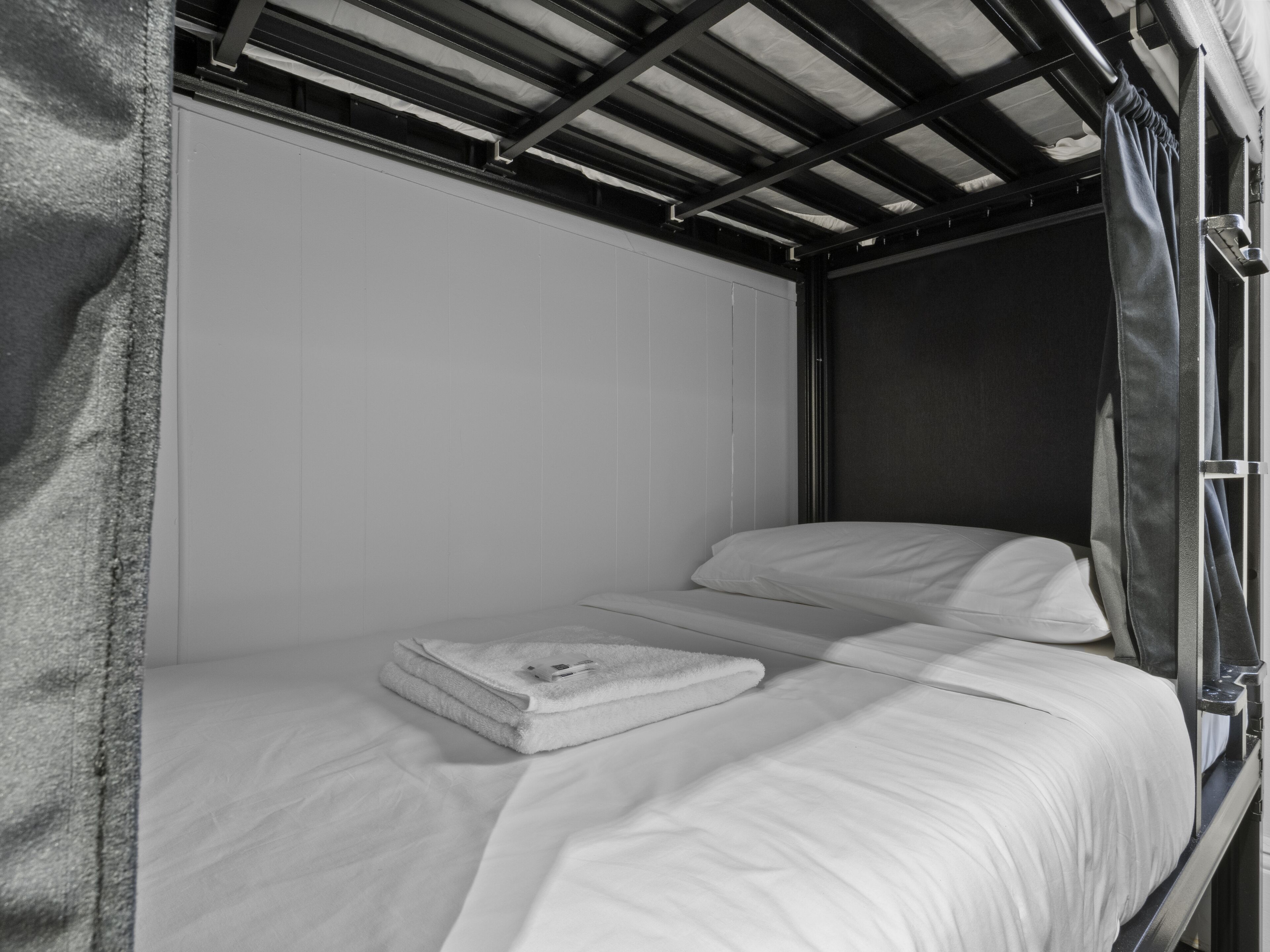 A single bed with white bedding and a folded towel, a metal frame, and a window with a curtain.