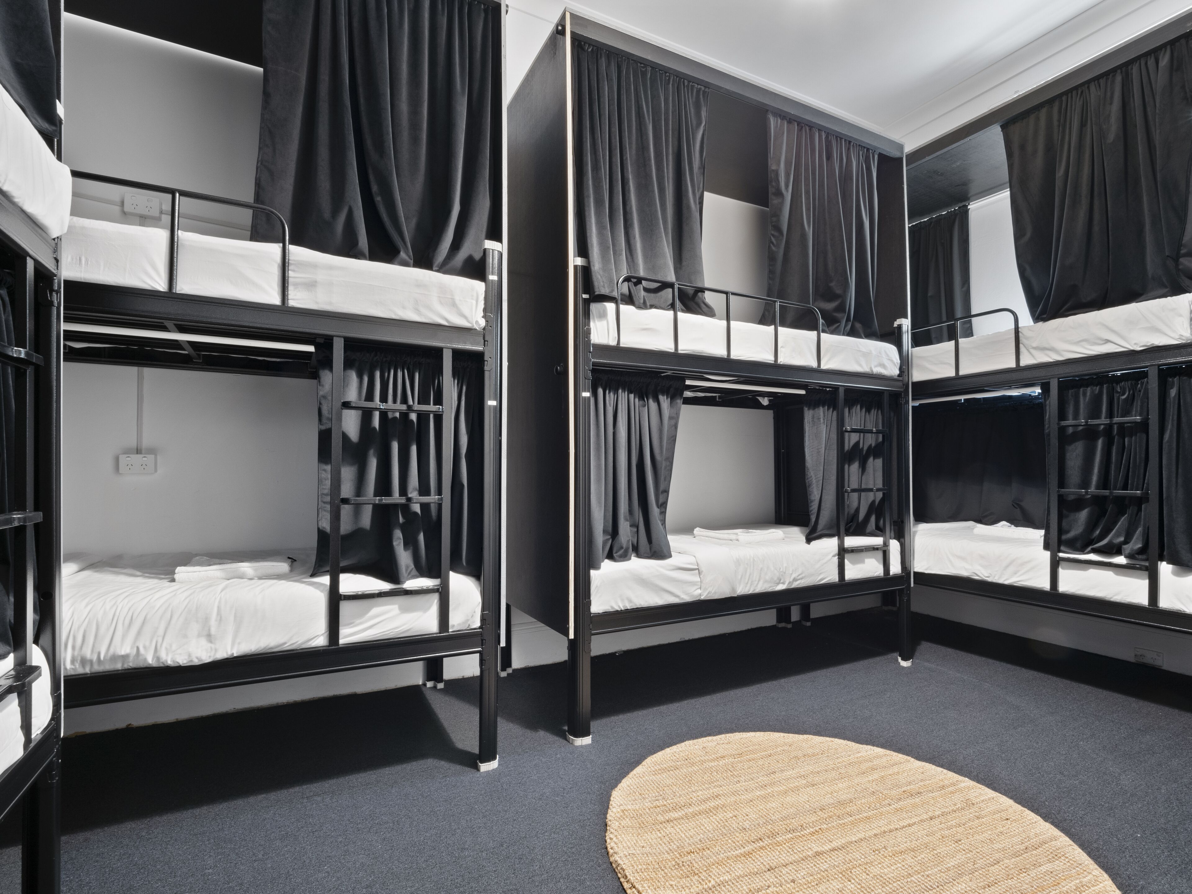 A bunk room with three bunk beds, each with curtains, a round rug, and a power outlet on the wall.