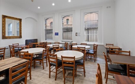 A dining area with round tables and wooden chairs, large windows with metal bars, and a white ceiling with recessed lighting.