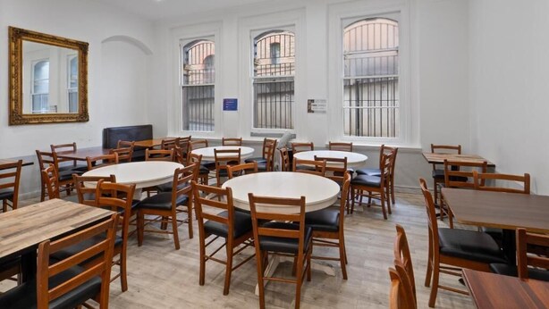 A dining area with round tables and wooden chairs, large windows with metal bars, and a white ceiling with recessed lighting. - Azzurro Pod Hotel Darling Harbour (Pyrmont)