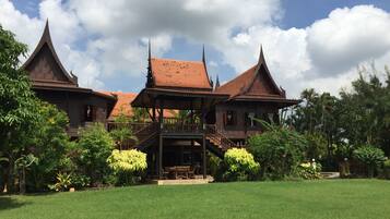 A traditional wooden building with multiple roofs and a central open pavilion, surrounded by lush greenery and a well-maintained lawn.