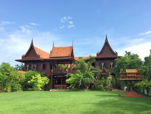 A traditional wooden building with multiple roofs and a central balcony, surrounded by lush greenery. - The Thai House Homestay (Bang Yai)