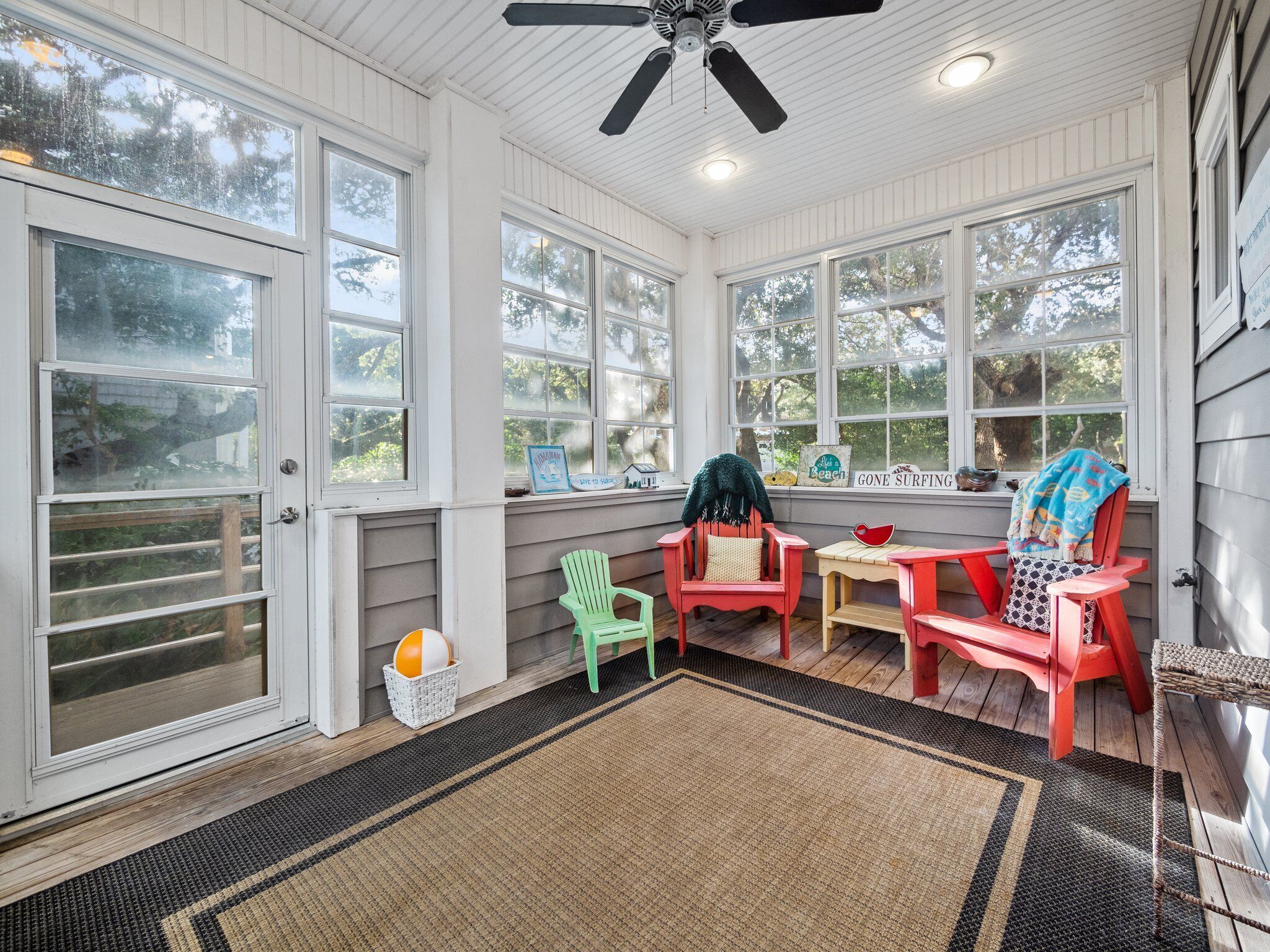 A sunroom with large windows, a ceiling fan, and colorful Adirondack chairs.