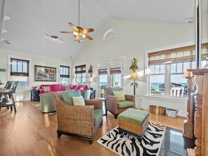 A spacious living room with wicker furniture, a ceiling fan, and large windows offering a view of the sea.