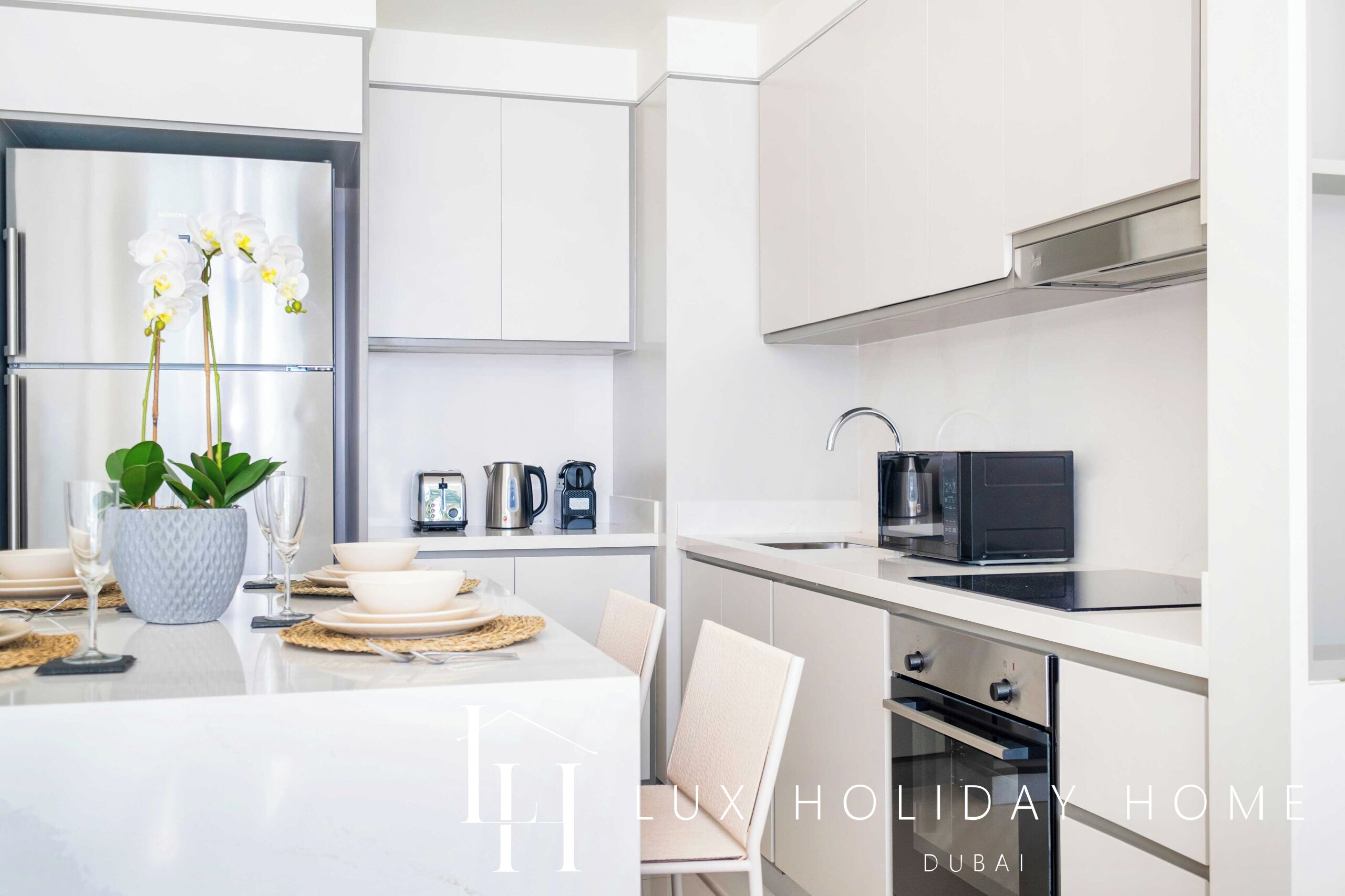 A modern kitchen with white cabinetry, a stainless steel refrigerator, a microwave, and an oven. There is a dining area with a table set for two, a potted plant, and a vase with flowers.