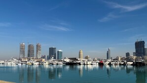 Un port de plaisance où sont amarrés des yachts, un ciel bleu azur et, au loin, des immeubles modernes de grande hauteur.