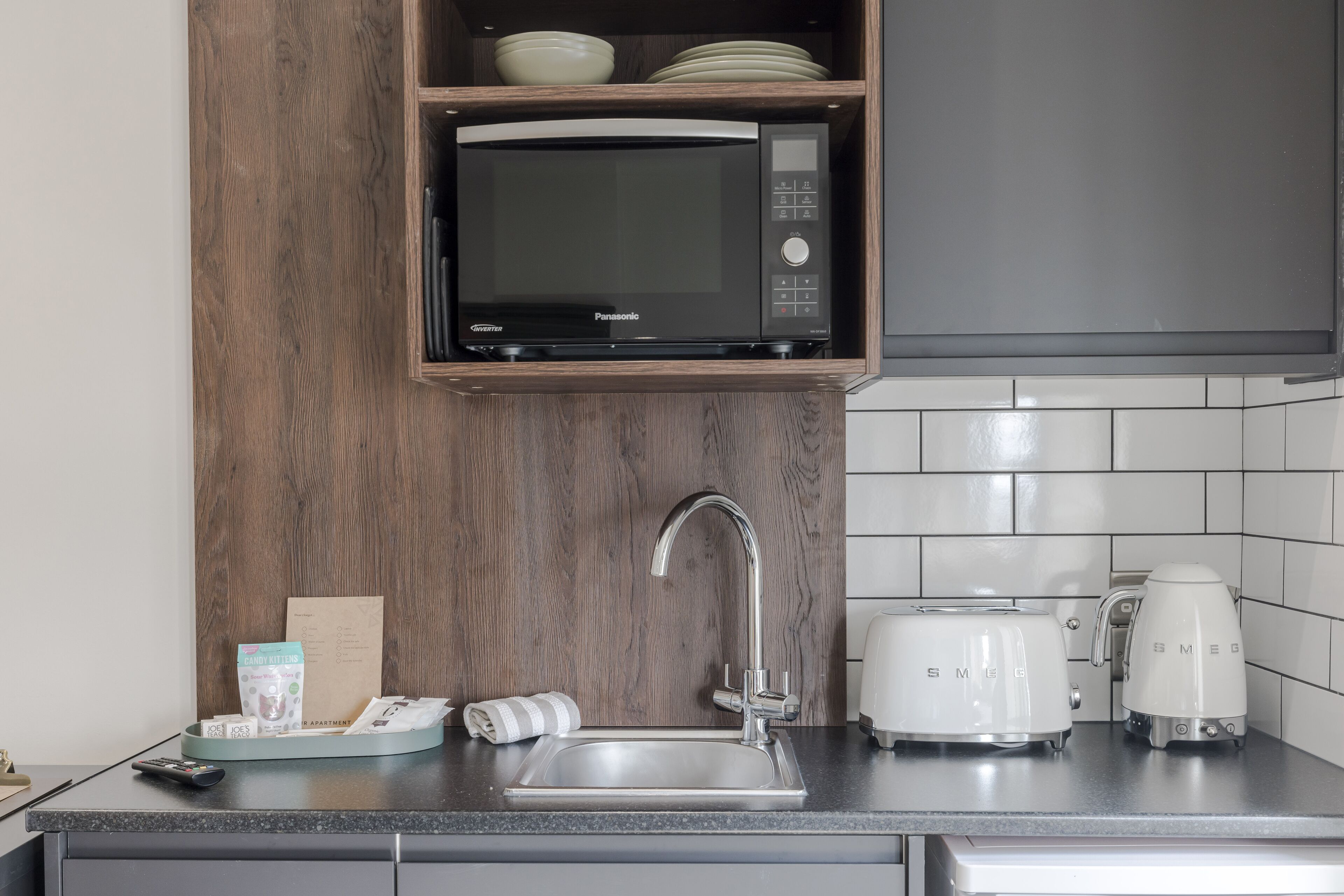 A kitchen with a microwave, toaster, and coffee maker on the countertop, a sink, and a wooden shelf above the microwave.