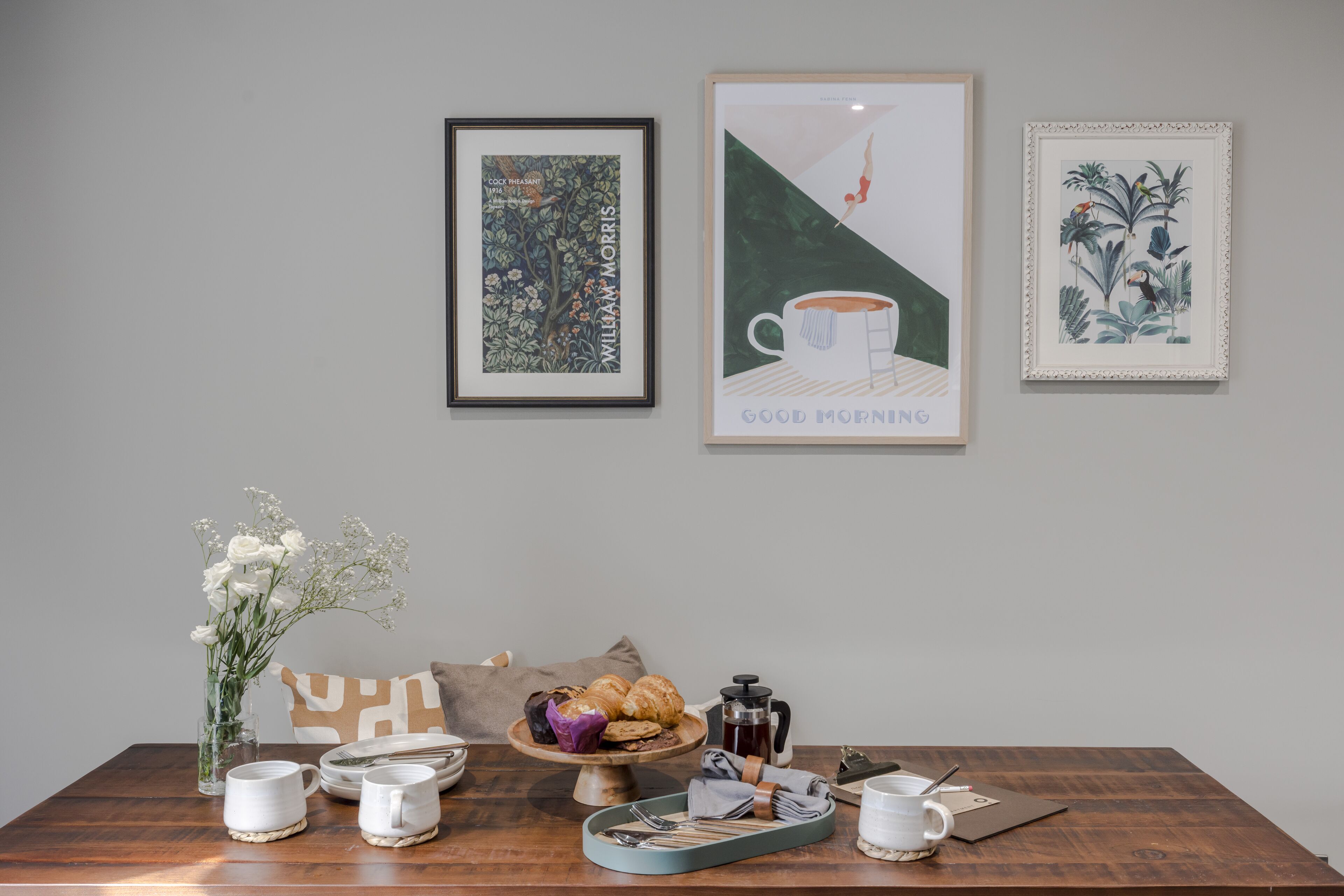 Uma mesa de jantar com um coador de café, bule, xícaras e uma bandeja com utensílios. Há obras de arte emolduradas na parede acima da mesa.