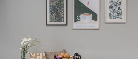 Uma mesa de jantar com um coador de café, bule, xícaras e uma bandeja com utensílios. Há obras de arte emolduradas na parede acima da mesa.