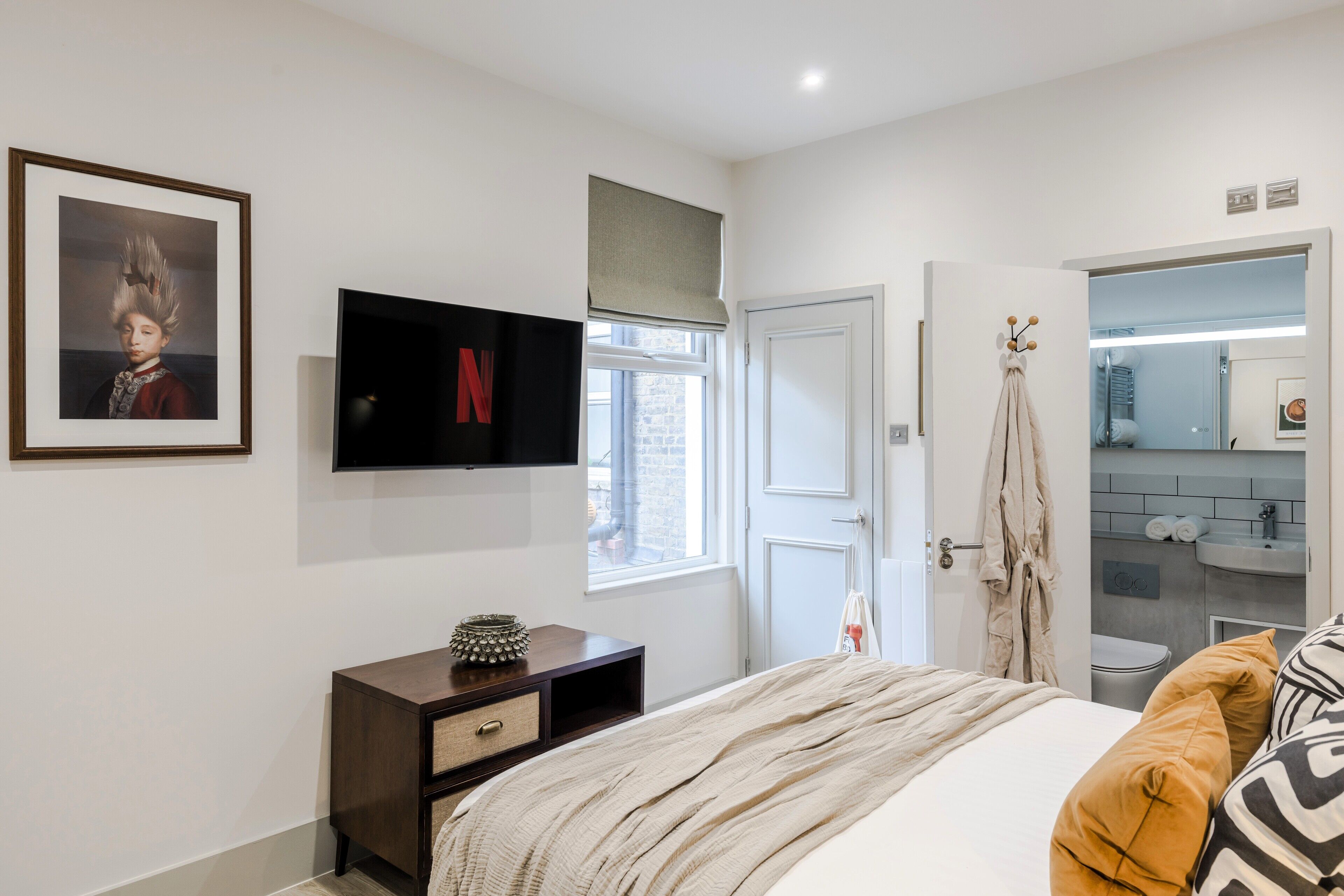 A modern bedroom with a bed, a TV mounted on the wall, a small cabinet, and a window with a view of the outside.