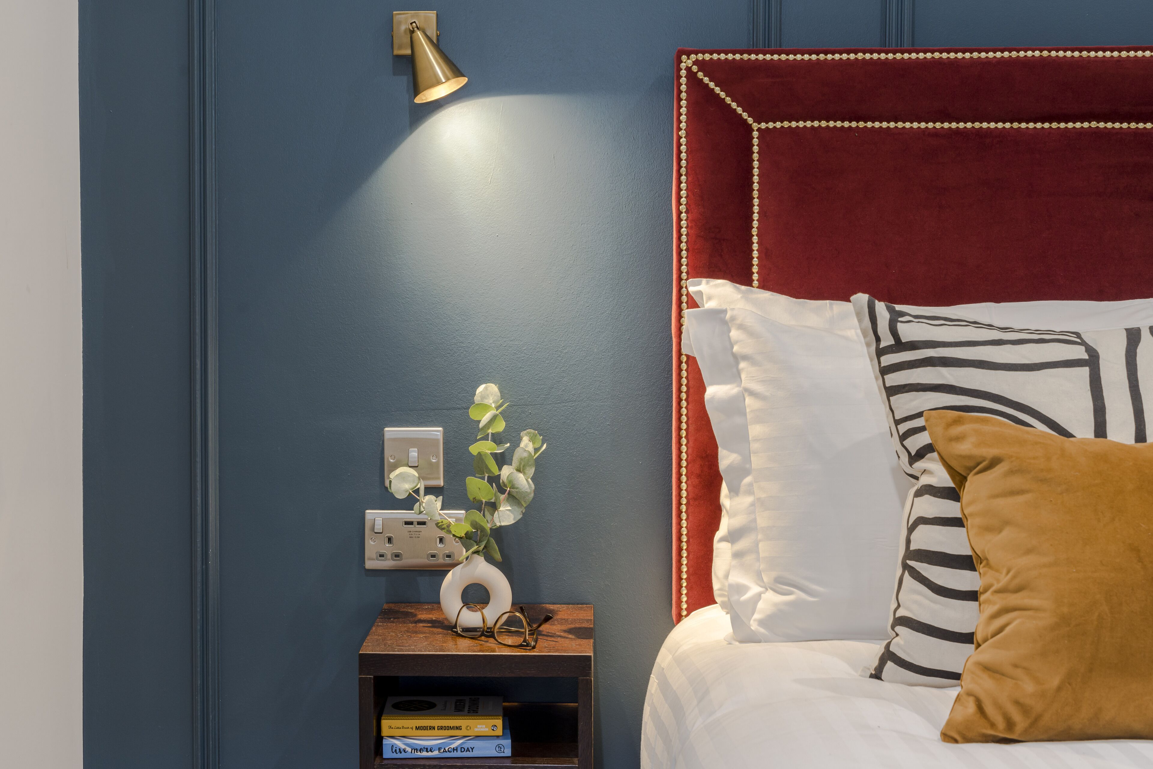 Un dormitorio con un cabecero tapizado en rojo, almohadas blancas y estampadas, una mesita de noche con un florero y una lámpara de pared.